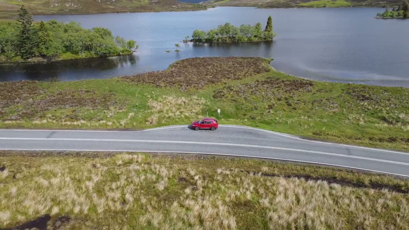 Aerial view of a red car on a winding highland road beside a loch